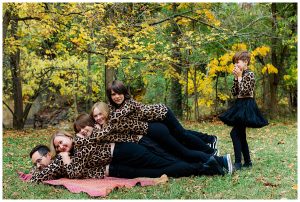 Family piles on top of each other for an awkward family photo with the fourth and youngest child standing to side, not sure how to get on top.