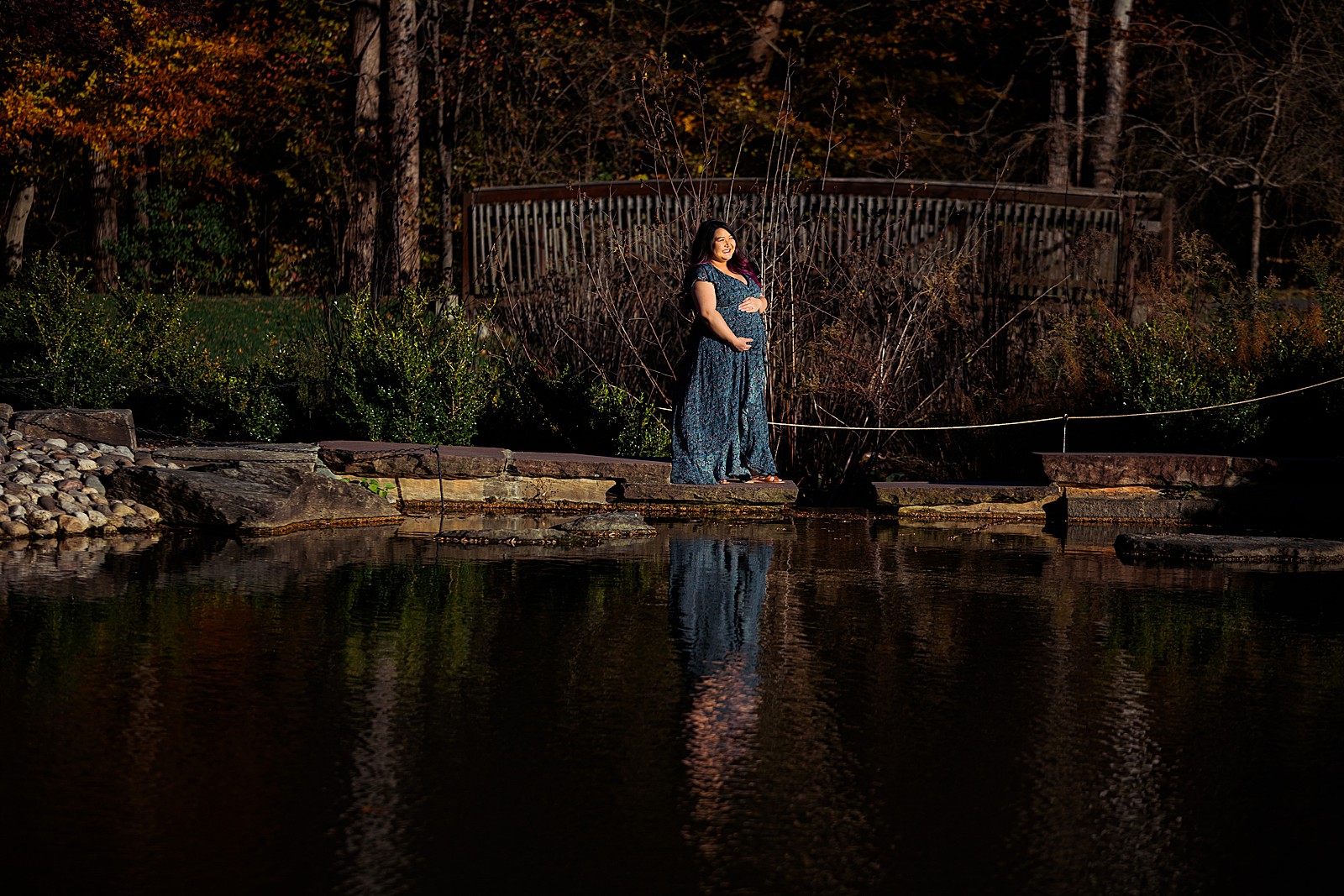 A woman stands across the lake holding her pregnant belly at her brookside gardens maternity session.