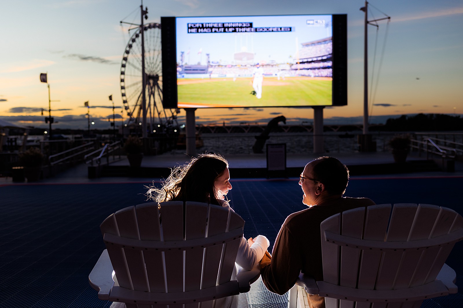 Bride and groom to be watch a baseball game outside on a big screen at the National Harbor. 