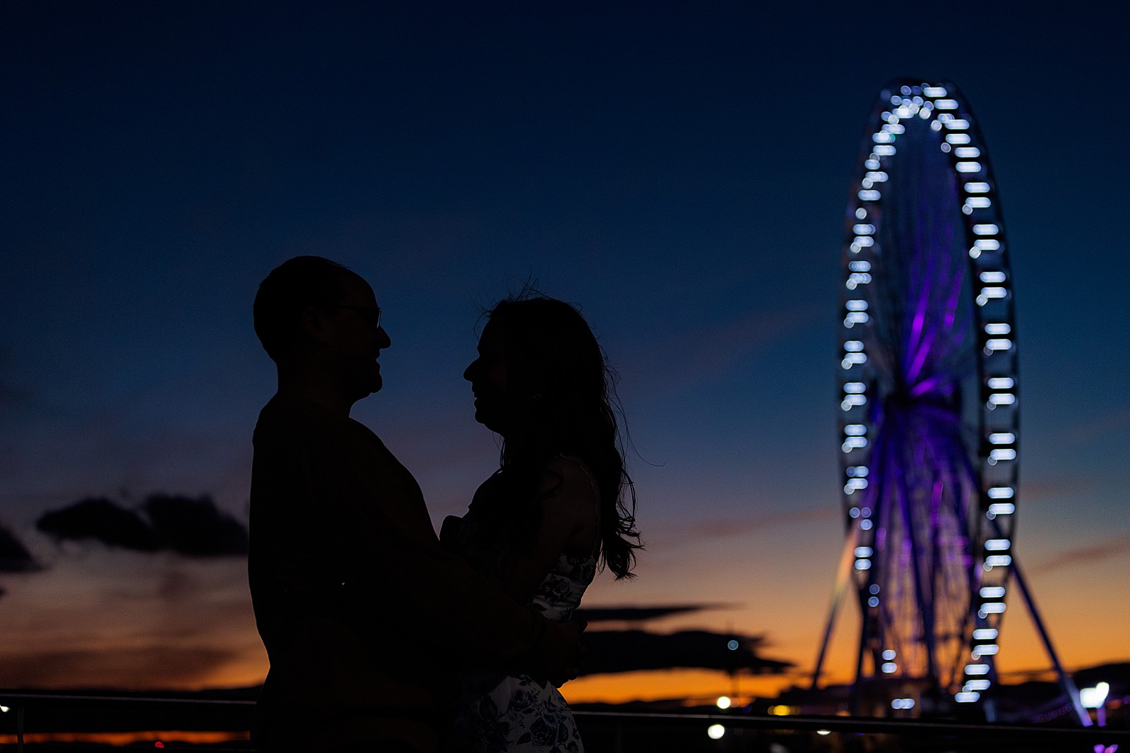 A couple is silhouetted against a twilight sky and a ferris wheel in the background. 