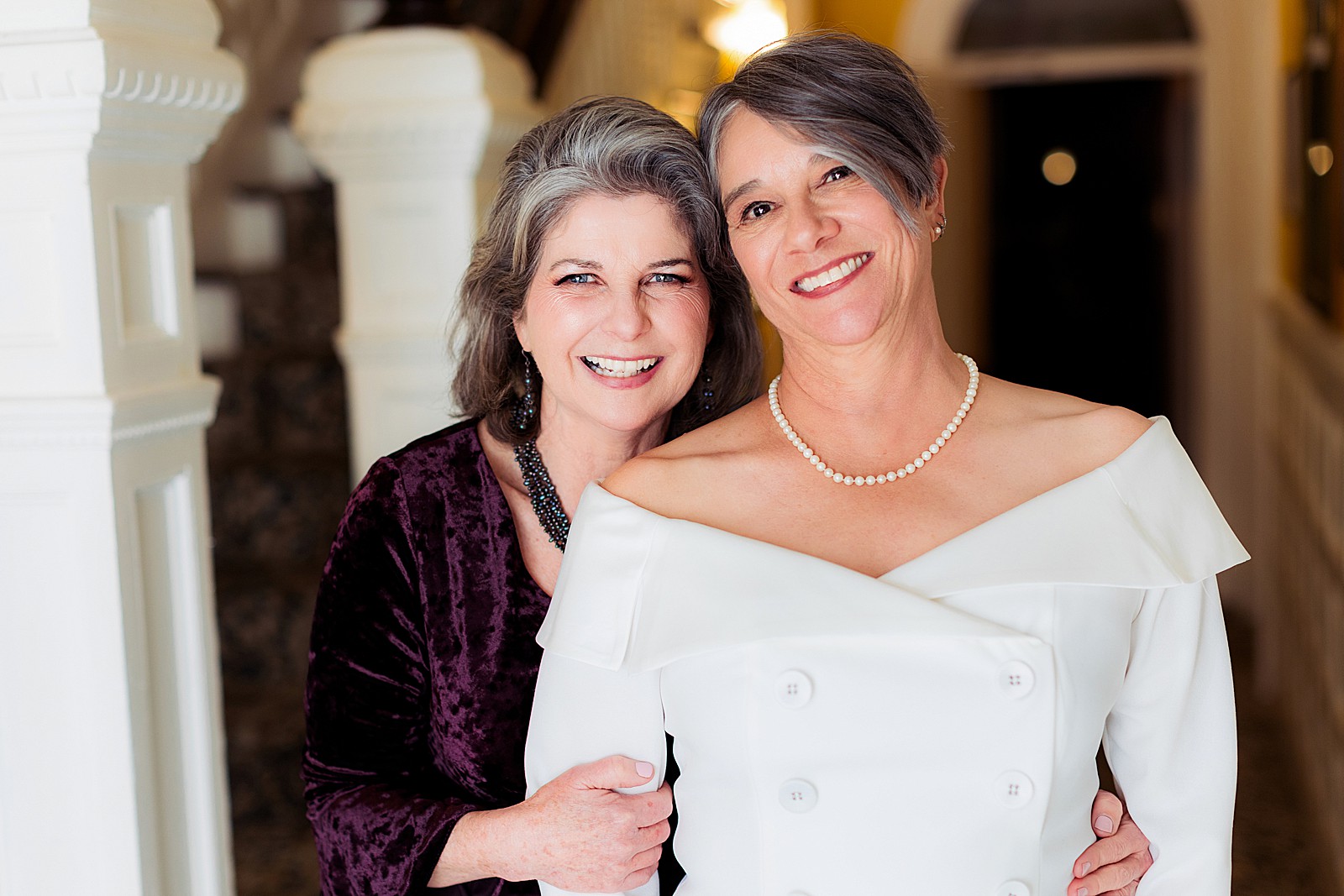 LGBTQ+ couple smiles at the camera linking arms in wedding attire.